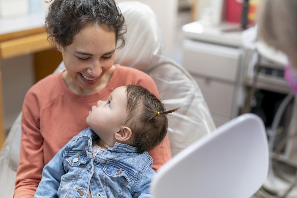 A loving mixed race mother and one year old daughter smile and look at each other while at a pediatric dental appointment. The woman is sitting in a dentist's chair in an examination room and is holding her daughter in her lap. The dentist is holding a mirror so the child can look at herself.