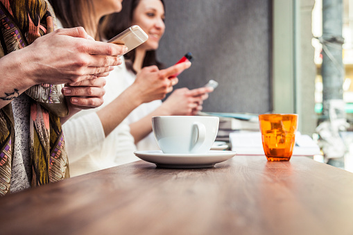Friends enjoy together an hot coffee cup in a bar in Copenhagen. Close up on the women hands using mobile phones.