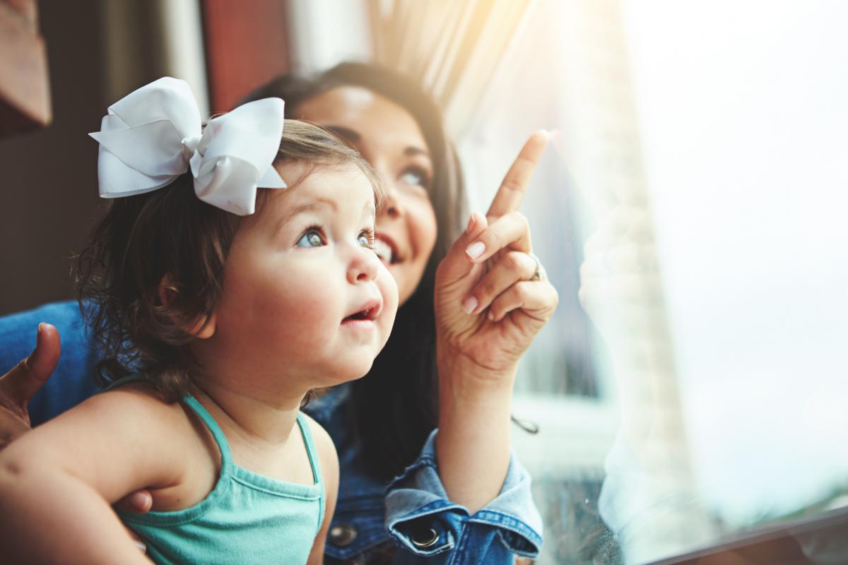 mother standing with toddler daughter, pointing and looking out a window