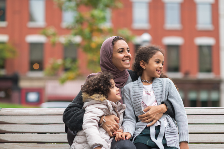 A mom and 2 daughters sitting on a bench in an urban area