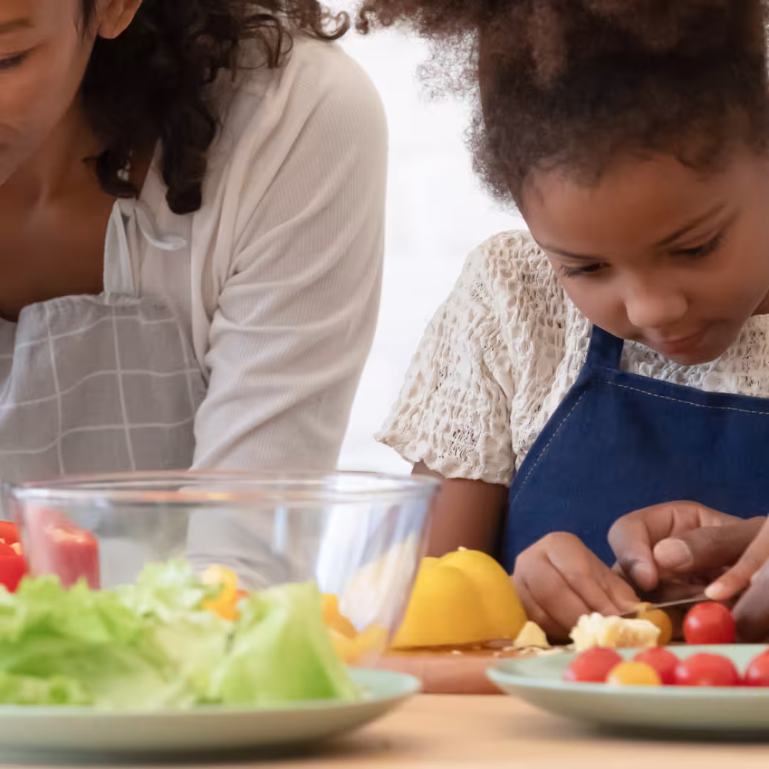 Happy family of four in the kitchen preparing food together. Parents are showing the two young sibling to slice fresh vegetables, peppers, tomatoes for salad on kitchen table or counter.