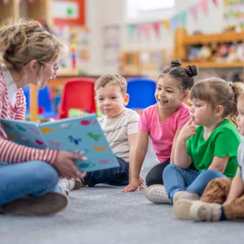 A preschool teacher sits on the floor of her classroom with a small group of students 