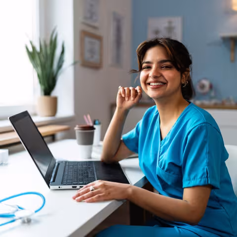 Portrait of a female doctor or a nurse of Indian ethnicity in a blue medical scrubs, using laptop at the modern ordination