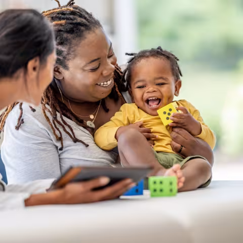 A young Mother brings in her toddler for a check-up. She is holding the little girl gently on the exam table as she talks with the doctor who is holding out a tablet with test results on it.