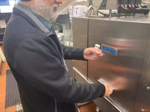 A public health inspector reads a test strip in front of a dishwasher in a restaurant kitchen.
