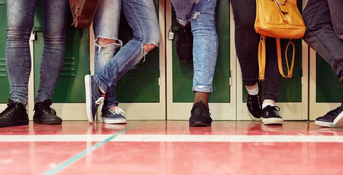 5 students standing in a row in front of lockers. Only legs and feet are shown