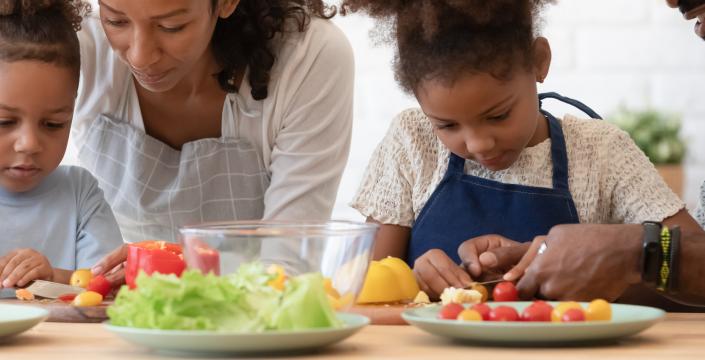 Happy family of four in the kitchen preparing food together. Parents are showing the two young sibling to slice fresh vegetables, peppers, tomatoes for salad on kitchen table or counter.