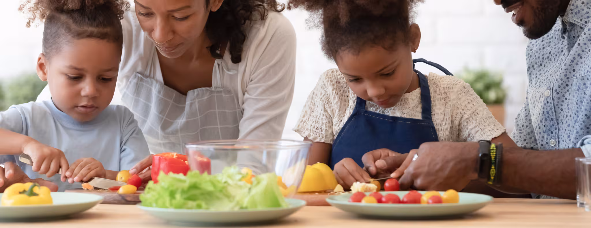 Happy family of four in the kitchen preparing food together. Parents are showing the two young sibling to slice fresh vegetables, peppers, tomatoes for salad on kitchen table or counter.