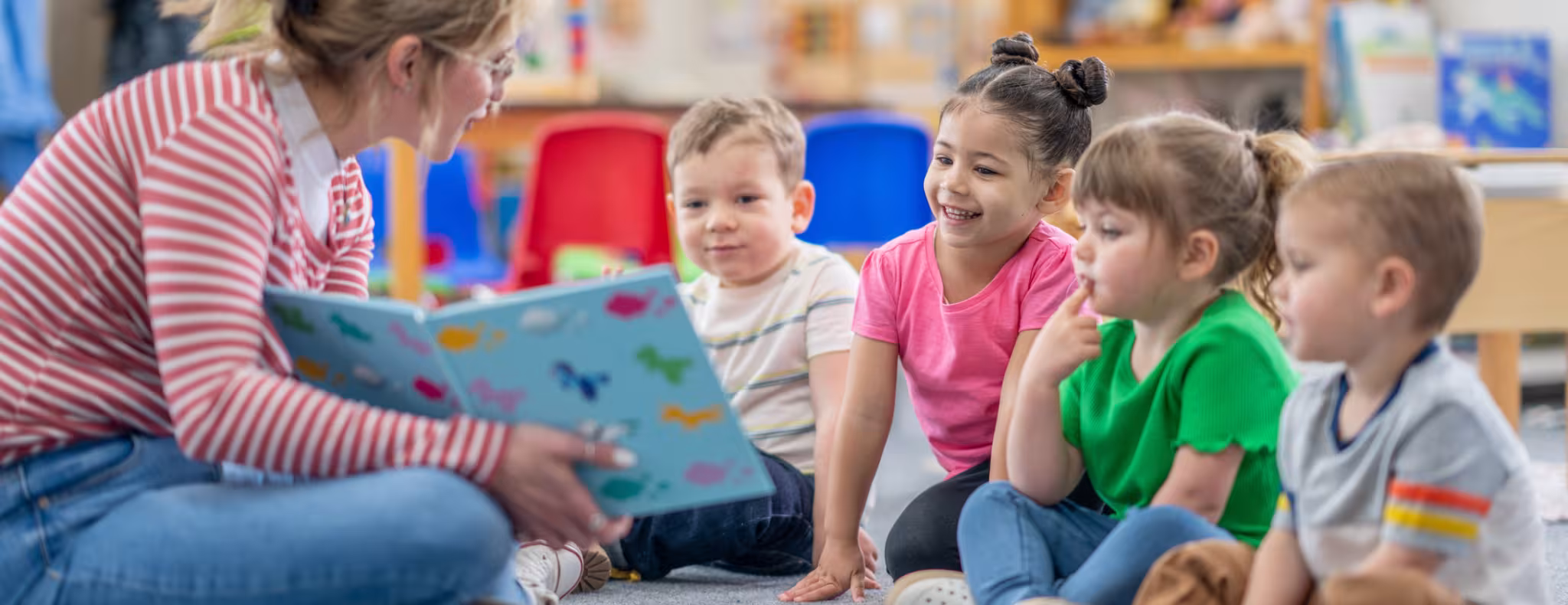 A preschool teacher sits on the floor of her classroom with a small group of students 