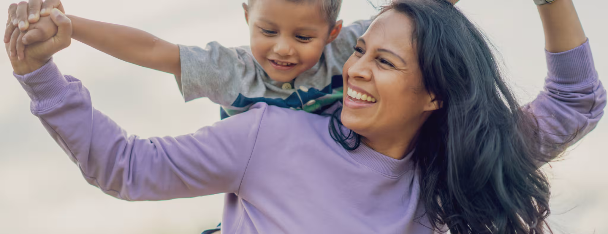 A Mother holds her son on her back and his arms out wide, as she pretends to fly with him. They are both dressed casually and are smiling as they pose for a portrait outside.