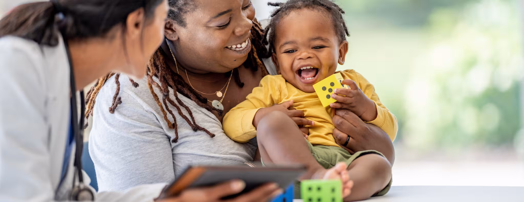 A young Mother brings in her toddler for a check-up. She is holding the little girl gently on the exam table as she talks with the doctor who is holding out a tablet with test results on it.