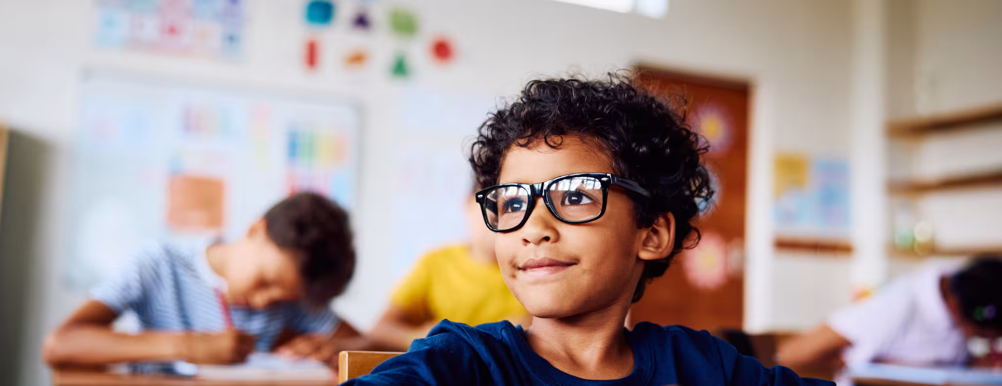 Schoolboy wearing eyeglasses sitting at desk, looking sideways, smiling in class