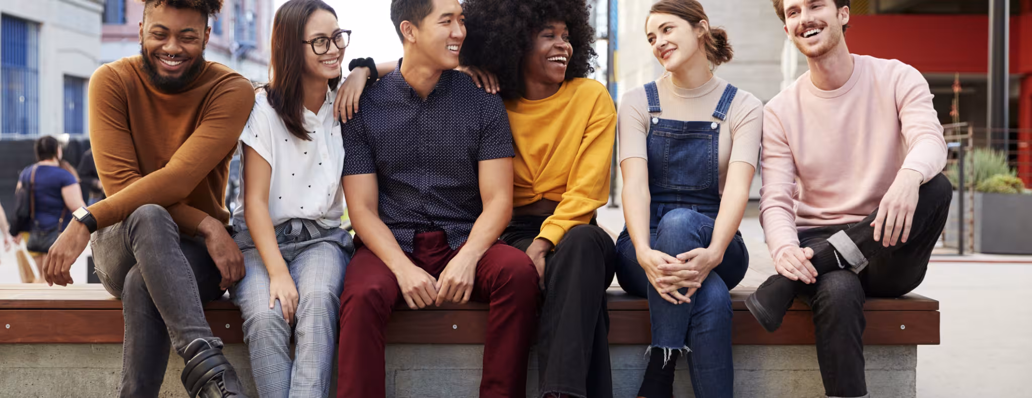 Six young adult friends sitting in a row on a bench in the street 