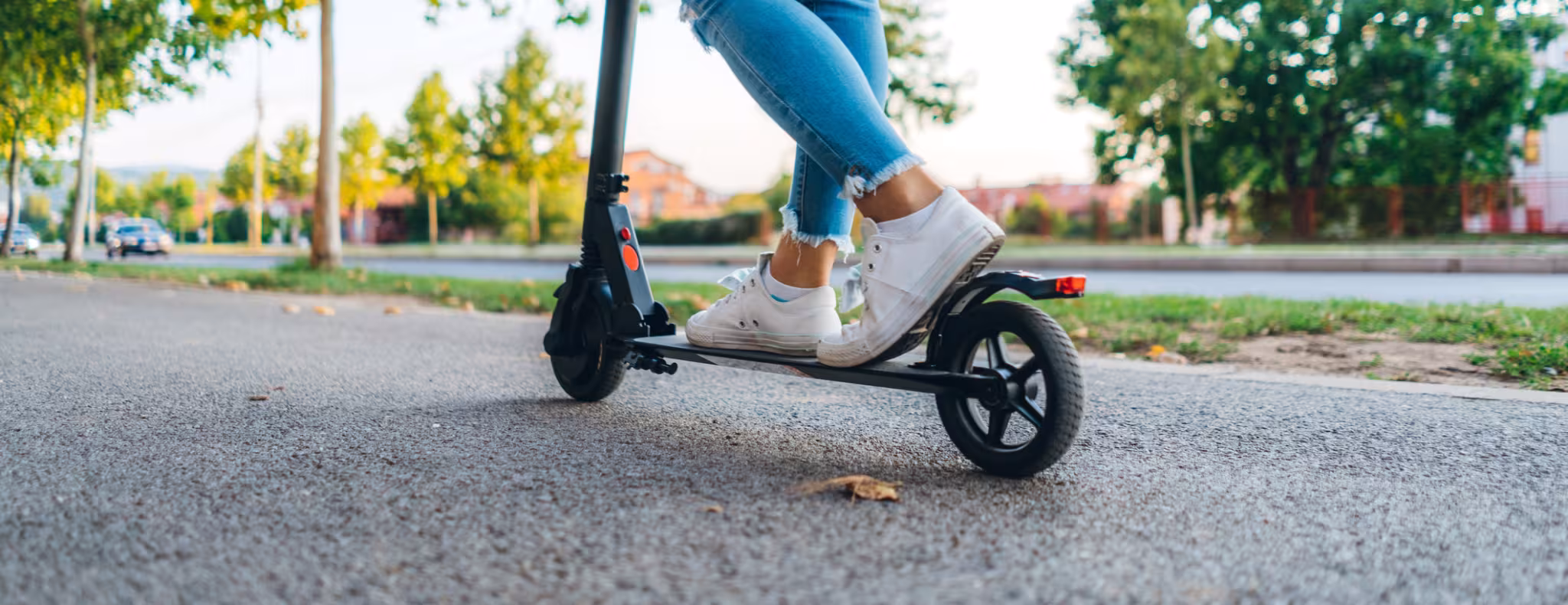 A close up of a pair of legs standing on an electric kick scooter on the pavement wearing jeans and sneakers on a sunny day.